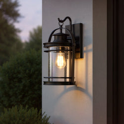 The Urban Ambiance UQL1431 Nautical Outdoor Wall Light (15"H x 9"W, Black Silk Finish, Cannes Collection) features an exposed bulb and glass shade, casting vintage lantern-style light at dusk against a white wall near bushes.