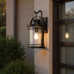 The Urban Ambiance UQL1432 Nautical Outdoor Wall Light from the Cannes Collection (18.25"H x 10.75"W, Black Silk Finish) glows warmly beside a garage door at dusk, surrounded by greenery.