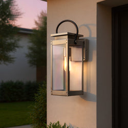 The Urban Ambiance UHP1132 Modern Farmhouse Outdoor Wall Light from the Darwin Collection features a stainless steel finish and glass, and is shown mounted by a door at dusk, illuminating the exterior with a lit bulb.