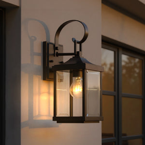 The Urban Ambiance UHP1192 Colonial Outdoor Wall Light from the Calderdale Collection features an Olde Bronze finish and clear glass panels, glowing beside a window on a beige exterior wall at dusk. Size: 15-1/8" x 5-1/2".