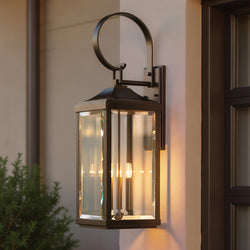 The UHP1194 Colonial Outdoor Wall Light by Urban Ambiance, in Olde Bronze, is mounted near a window and green shrubbery on a beige wall.