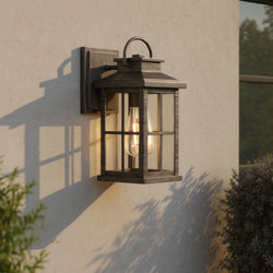 The Urban Ambiance UHP1294 Transitional Outdoor Wall Sconce (12''H x 5''W) in Aged Pewter from the Anaheim Collection is mounted on a light exterior wall near green foliage, delivering timeless farmhouse-style outdoor lighting.