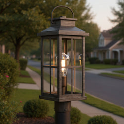 The Urban Ambiance UHP1297 Anaheim Collection Outdoor Post Light, 20.25''H x 7''W in aged pewter, features powder-coated aluminum and clear glass panes with a visible bulb—perfect for suburban streetscapes with houses and greenery.