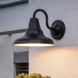 The Urban Ambiance UHP1363 Industrial Outdoor Wall Sconce (10.5''H x 12''W, Midnight Black, Pittsburgh Collection) is mounted on a light stucco wall, casting a warm downward glow with its stylish gooseneck arm design.