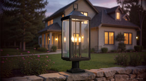 The UHP1400 Farmhouse Outdoor Post Light from Urban Ambiance’s Calderdale Collection, featuring a Midnight Black finish and measuring 26.875′‘ high by 9.5’’ wide, sits atop a stone wall at dusk in front of a home. Its lantern glows gently behind beveled glass panes, casting warm light over the yard and nearby windows to create an inviting ambiance.