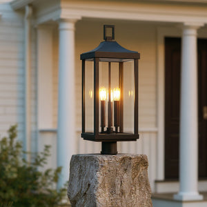 The Urban Ambiance UHP1400 Farmhouse Outdoor Post Light, 26.875''H x 9.5''W in a midnight black finish from the Calderdale Collection, is shown mounted on a stone pillar and illuminated in front of a house with white columns.