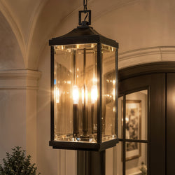 A striking Urban Ambiance UHP1401 Farmhouse Outdoor Pendant from the Calderdale Collection, featuring a midnight black finish and elegant beveled glass panels in a rectangular silhouette reminiscent of a colonial lantern, hangs from the ceiling of an entryway with arched details, its three visible bulbs casting warm illumination.