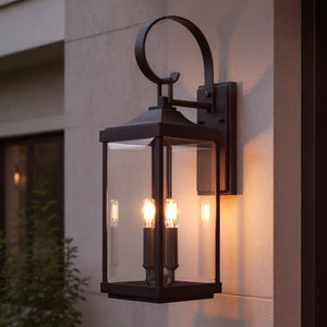 The Urban Ambiance UHP1403 Calderdale Collection Transitional Outdoor Wall Sconce (21.75''H x 7''W) in Midnight Black adds Colonial-inspired elegance with three exposed bulbs and clear beveled glass, glowing beautifully on a light exterior wall at dusk.