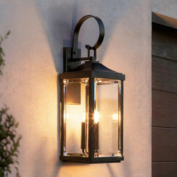 The UHP1404 Transitional Outdoor Wall Sconce from the Calderdale Collection by Urban Ambiance features a glowing bulb and a sleek midnight black finish, mounted on a light-colored stucco wall near a wooden door.
