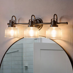 An Urban Ambiance UHP2044 Luxe Industrial Bath Fixture from the Harlow Collection, featuring a Fashion Bronze finish, is mounted above a round mirror in a bathroom with a white door and tiled wall.