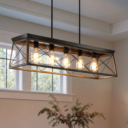 The UHP2126 Luxe Industrial Linear Chandelier by Urban Ambiance, featuring an Olde Bronze finish from the Berkeley Collection and measuring 9″ high by 38″ wide, hangs from the ceiling in a room with large windows and a plant in the foreground, creating a striking piece of rustic lighting with its rectangular metal-and-wood design and four exposed bulbs.