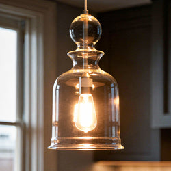 An Urban Ambiance UHP3043 Modern Farmhouse Pendant Light from the Dundee Collection, featuring a clear hand-blown glass shade and a brushed nickel finish, hangs from the ceiling near a window in a room with dark walls.