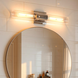 The Urban Ambiance UHP4052 Cosmopolitan Bath Light from the Tustin Collection features a minimalist, wall-mounted design with two exposed bulbs positioned above a round mirror, reflecting portions of a tiled bathroom wall and countertop adorned with various toiletries. The polished chrome finish enhances the modern appeal of this 5′‘H x 20.375’’W fixture.