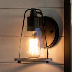 The UHP4074 Coastal Wall Sconce by Urban Ambiance, from the Kempsey Collection, features a midnight black finish and clear glass shade, bringing industrial style with coastal charm to light-colored wood panel walls.