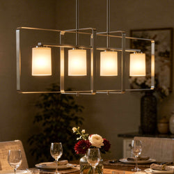 A UHP4101 Modern Chandelier from the Broome Collection by Urban Ambiance, featuring a brushed nickel finish and four sculptural frosted glass shades, hangs above a dining table set with plates, glasses, and a flower centerpiece in a warmly lit dining room.