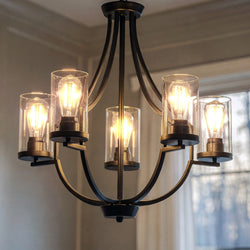 The Urban Ambiance UHP4369 Contemporary Chandelier (26''H x 26''W, Midnight Black, Mesa Collection) with five exposed bulbs and cylindrical glass shades hangs from the ceiling in a room with light walls and a window.
