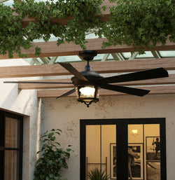 The Urban Ambiance UHP9181 Catalina ceiling fan, 19.5"H x 52"W in black iron, adds a modern farmhouse touch to a wooden pergola above a patio with glass doors, framed pictures inside, and lush green plants nearby.
