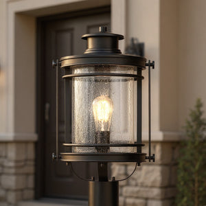 A close-up of the Urban Ambiance UQL1010 Cannes Collection Outdoor Post Light in Black Silk Finish, 19.5"H x 10.5"W, featuring seeded glass panes and a lit bulb, standing at a house entrance.