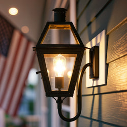 The Urban Ambiance UQL10193 English Country Outdoor Wall Sconce in Earth Black glows against house siding at dusk, lantern-style, with an American flag waving in the background. Marine-grade and from the Paris Collection.