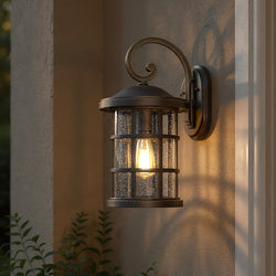 An Urban Ambiance UQL1045 Craftsman Outdoor Wall Light from the Vienna Collection, 17.75"H x 10"W with a Parisian Bronze finish, is mounted on a stucco wall near green foliage.