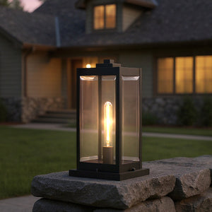 The Urban Ambiance UQL1336 Modern Farmhouse Outdoor Pier Light, 16"H x 7.5"W in a natural black finish from the Quincy Collection, sits on a stone wall, its glowing bulb casting warmth as dusk settles over the house and lawn.