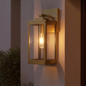 The Urban Ambiance UQL1340 Quincy Collection Farmhouse Outdoor Wall Light, 14"H x 5"W in Antique Brass, is mounted on a beige wall near foliage.