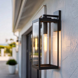 The UQL1352 Modern Farmhouse Outdoor Wall Light by Urban Ambiance, from the Quincy Collection, features a sleek rectangular design with visible bulb in estate bronze, and is shown illuminated on an exterior wall during daylight.
