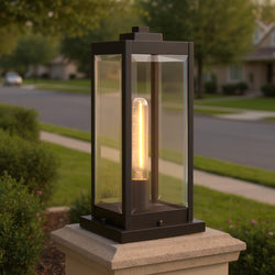 The Urban Ambiance UQL1355 Modern Farmhouse Outdoor Pier Light from the Quincy Collection, in Estate Bronze and measuring 16"H x 7.5"W, features clear glass panels and a lit bulb, mounted on a stone pillar beside a suburban street.
