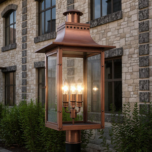 The Urban Ambiance UQL1410 Vintage Outdoor Post/Pier Light, 26"H x 12"W in Rustic Copper from the Longview Collection features four glowing bulbs, adding timeless charm to a stone building with arched windows and greenery.