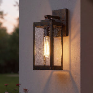 The UQL1632 Modern Farmhouse Outdoor Wall Sconce (14.25''H x 5''W) in Bygone Bronze from Urban Ambiance's Quincy Collection features clear glass and a visible bulb, mounted on stucco and glowing beautifully at dusk.