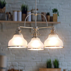 The Urban Ambiance UQL2330 Industrial Chandelier from the Pasadena Collection, featuring a brushed nickel finish and three opal etched glass shades, hangs lit in a room with a white brick wall and plant-filled shelves.