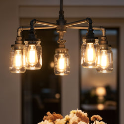 The UQL2663 Industrial Chandelier from Urban Ambiance's Dallas Collection, featuring an antique black finish and measuring 23 inches high by 26 inches wide, hangs above the table with a floral centerpiece. This distinctive farmhouse fixture fills the room with warm light, infusing the space with rustic industrial charm.