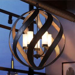The Urban Ambiance UQL2782 Rustic Chandelier from the Walsall Collection, featuring a weathered black finish, hangs from the ceiling with white cylindrical lampshades in front of a window at dusk.