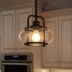 An Urban Ambiance UQL3331 Utilitarian Pendant Light from the Clearwater Collection, featuring a clear glass shade and dark bronze finish, hangs in a kitchen with beige cabinets and a built-in oven.