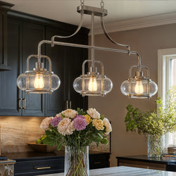 Three UQL3380 Utilitarian Chandeliers (Clearwater Collection, Urban Ambiance) with exposed bulbs in brushed nickel hang above a kitchen island with flowers; dark cabinets and greenery finish this modern farmhouse look.