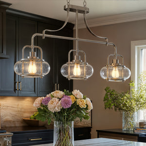 Three UQL3380 Utilitarian Chandeliers (Clearwater Collection, Urban Ambiance) with exposed bulbs in brushed nickel hang above a kitchen island with flowers; dark cabinets and greenery finish this modern farmhouse look.