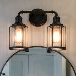 The UQL3502 Luxe Industrial Bath Vanity Light by Urban Ambiance, a black metal dual sconce with clear glass covers, is mounted above a round mirror on a light-colored wall.