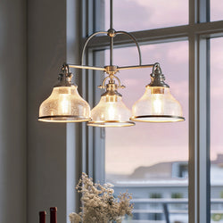 The Urban Ambiance UQL3667 Traditional Chandelier from the Pasadena Collection, featuring a brushed nickel finish and three clear glass shades, hangs before a large window at dusk, with white flowers and bottles arranged below.