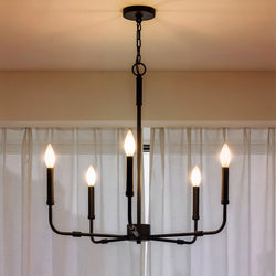 The Urban Ambiance UQL3803 Modern Farmhouse Chandelier, 25.25''H x 24''W with a matte black finish from the Bideford Collection, features five candle-style bulbs and hangs in front of sheer white curtains.