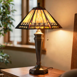 The Urban Ambiance UQL7018 Copeland Tiffany Table Lamp, measuring 23.5''H x 16''W with a russet bronze base and stained glass in Mediterranean style, is lit on a wooden surface near a window with a plant in the background.