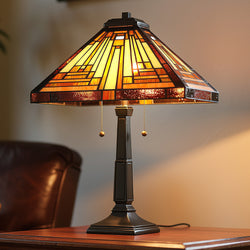 The Urban Ambiance UQL7180 Waltham Tiffany Table Lamp, 23''H x 16''W, features a vintage bronze finish and craftsman-style stained glass, glowing on a wooden side table beside a brown leather chair.