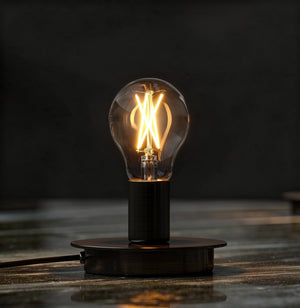 An Urban Ambiance UBB2000 Luxury LED A19 bulb, 60W equivalent, vintage Edison style with visible filament and E26 base, glows warmly on a dark round base atop a reflective surface against a dark background.