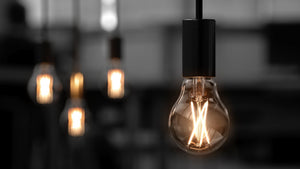Close-up of an Urban Ambiance UBB2000 Luxury LED Bulb, 60W equivalent, A19 with E26 base and 2700K warm white glow, hanging from the ceiling as other similar vintage Edison style bulbs softly blur in the background.