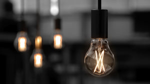A close-up of a lit Urban Ambiance UBB2001 Luxury LED Bulb (A19 shape, E26 base, 60W equivalent, 3000K soft white) with a visible filament, plus two softly glowing blurred bulbs in the background.