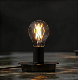An Urban Ambiance UBB2001 Luxury LED Bulb (60W Equivalent, Vintage Edison A19, E26 Base, 3000K soft white) glows atop a round black base on a reflective surface against a dark background.