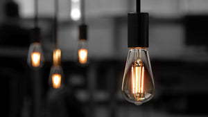 A close-up of an Urban Ambiance UBB2090 Luxury LED ST19 bulb (E26 base, 2700K warm white, 60W equivalent) glows in the foreground, while similar bulbs softly light the blurred background. Available in a 4-pack, vintage Edison style.