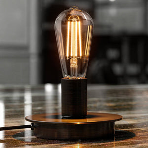 An Urban Ambiance UBB2090 Luxury LED Bulb, ST19 vintage Edison style with exposed filaments, emits a warm 2700K glow from its E26 base as it shines on a reflective surface; available in a 4-pack, 60W equivalent.