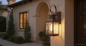 The Urban Ambiance UFS1000 Calderdale Collection 15''H x 5.5''W outdoor wall sconce in genuine copper and dark patina glows beside a wooden door on a beige stucco house with arched entryways and lit windows at dusk.
