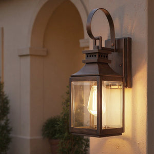 The Urban Ambiance UFS1000 Calderdale Collection 15''H x 5.5''W genuine copper outdoor wall sconce with dark-patina finish glows on the building's exterior, framed by plants and an archway in the background.