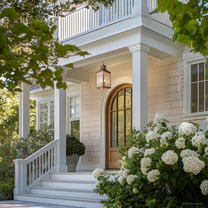 A front porch with white columns, a wood door with arched window, white stairs, potted plants and hydrangeas features the Urban Ambiance UFS1004 Calderdale Collection Outdoor Pendant Light in genuine copper with a dark-patina finish.